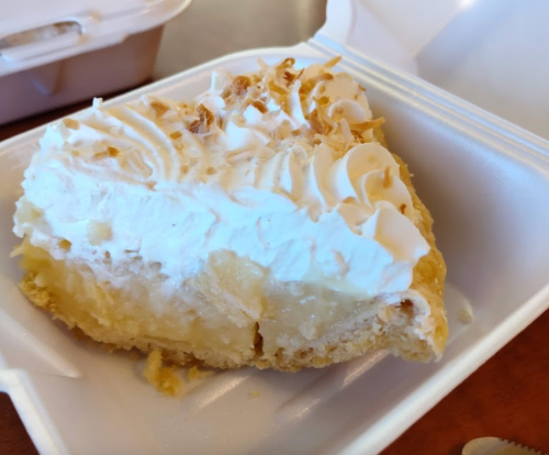 A slice of creamy pie topped with whipped cream and toasted coconut flakes, served in a takeout container.