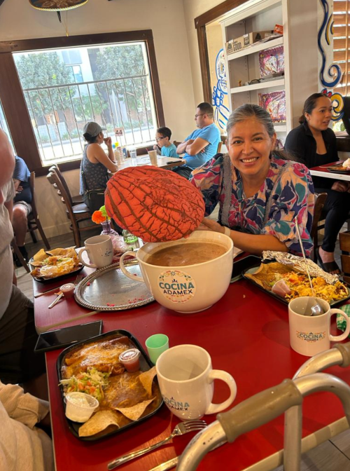A woman smiles while holding a large bowl of soup at a busy restaurant table with various dishes around her.