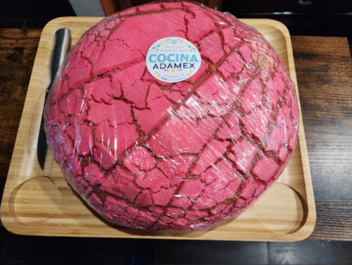 A round, pink cake wrapped in plastic, placed on a wooden tray with a knife beside it.