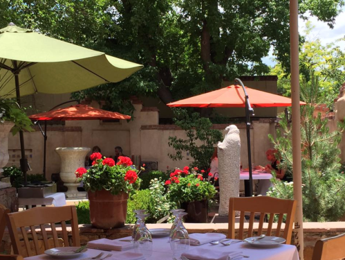A sunny outdoor dining area with green umbrellas, red flowers, and a stone sculpture, creating a serene atmosphere.