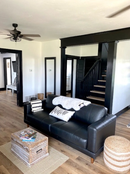 A cozy living room featuring a black sofa, decorative pillows, a wooden coffee table, and a staircase in the background.