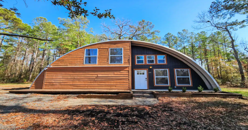 A modern, curved wooden house surrounded by trees, featuring large windows and a welcoming entrance.