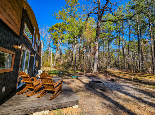 A cozy outdoor space with wooden chairs, a fire pit, and tall trees under a clear blue sky.