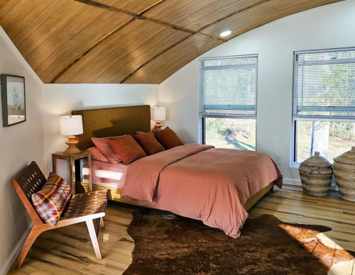 Cozy bedroom with a wooden ceiling, a pink bedspread, two lamps, a chair, and natural light from large windows.