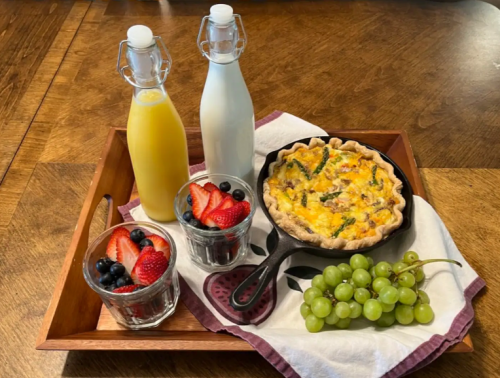 A wooden tray with a quiche, two bottles of juice, and glasses of berries, alongside green grapes.