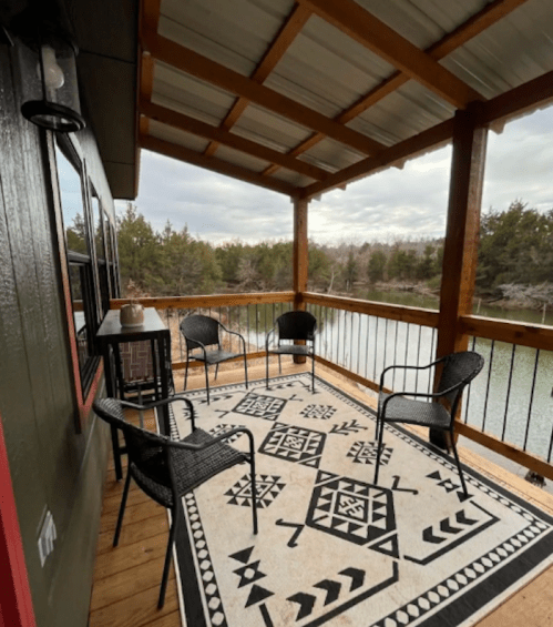 A cozy outdoor patio with four chairs and a patterned rug, overlooking a serene body of water and trees.