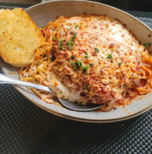 A bowl of cheesy baked spaghetti with marinara sauce, topped with herbs, alongside a slice of garlic bread.