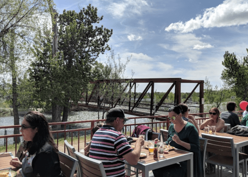 People dining outdoors on a patio with a view of a river and a bridge under a partly cloudy sky.