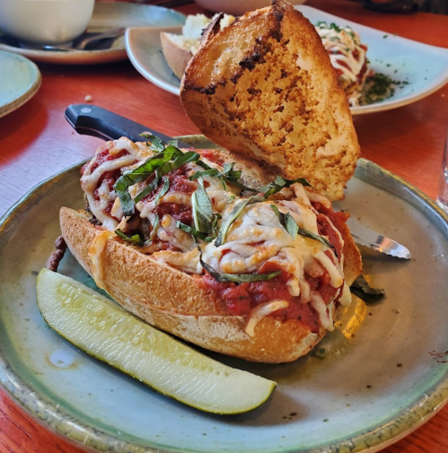 A sandwich with marinara sauce, melted cheese, and fresh basil on a plate, accompanied by a pickle slice.
