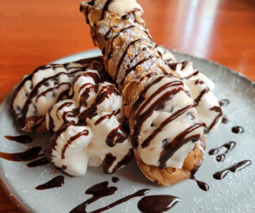 A plate of cannoli filled with cream and drizzled with chocolate sauce, garnished with a sprinkle of powdered sugar.