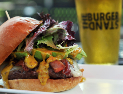 A close-up of a juicy burger with lettuce, cheese, and mustard, alongside a glass of beer.
