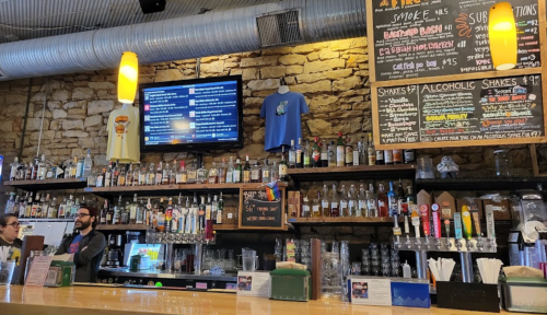 A bar with a stone wall, shelves of liquor, a menu board, and a TV displaying tweets. Two staff members are present.