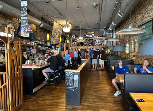 A lively restaurant interior with a bar, patrons seated at tables, and a rustic stone wall decor.