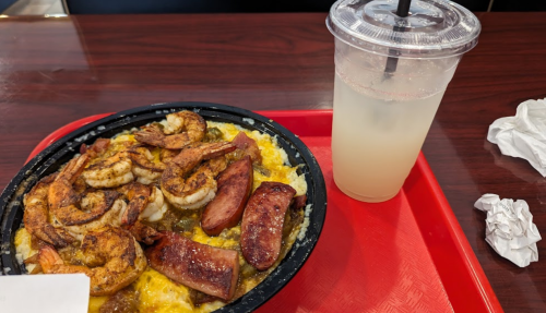 A bowl of shrimp, sausage, and rice next to a clear drink in a plastic cup on a red tray.