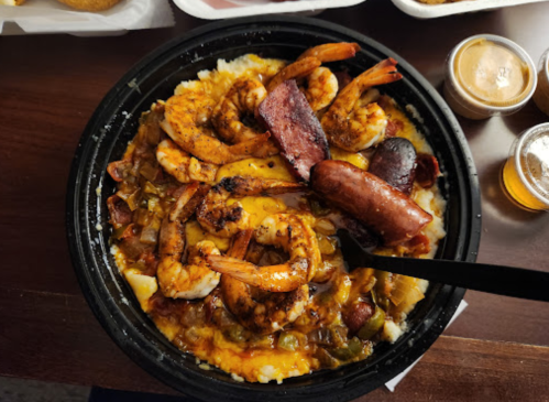 A bowl of shrimp, sausage, and cheese over a bed of grits, garnished with spices and served with sauces on the side.