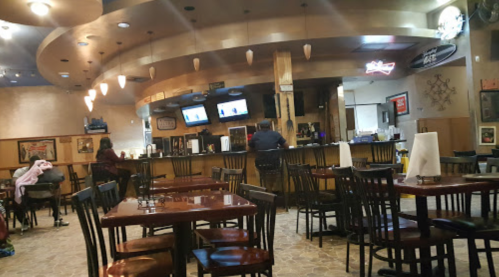 Interior of a restaurant with wooden tables and chairs, a bar area, and two TVs on the wall. A few patrons are seated.