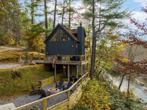 A modern cabin with a deck overlooking a river, surrounded by trees and autumn foliage. Cozy outdoor seating area.