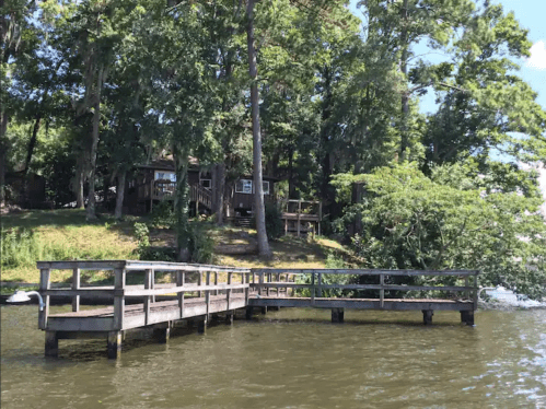 A wooden dock extends into a calm lake, surrounded by trees and a house on the shore in the background.