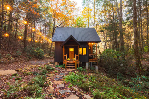 A cozy cabin in the woods, surrounded by autumn foliage and string lights, with a stone pathway leading to the entrance.