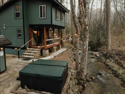 A cozy green cabin by a stream, featuring a hot tub and surrounded by trees in a serene outdoor setting.