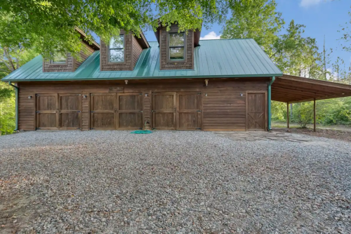 A wooden garage with a green metal roof, featuring three doors, surrounded by gravel and greenery.