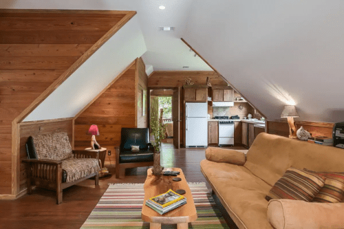 Cozy wooden living room with sloped ceilings, a sofa, chairs, and a kitchen visible in the background.
