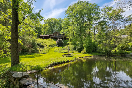 A serene landscape featuring a pond, lush greenery, and a picnic area with a house in the background under a clear blue sky.