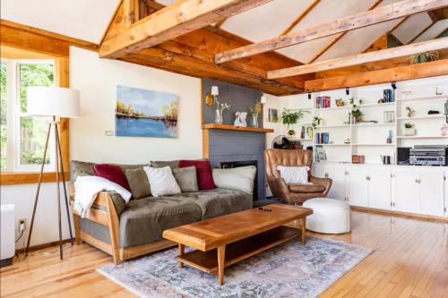 Cozy living room with wooden beams, a sofa, coffee table, and shelves filled with books and decor. Bright and inviting space.