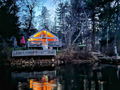A cozy cabin by the water, illuminated at dusk, surrounded by trees and reflecting in the calm lake.