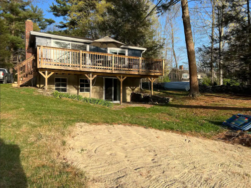 A house with a wooden deck sits on a grassy yard, surrounded by trees and a sandy area in the foreground.