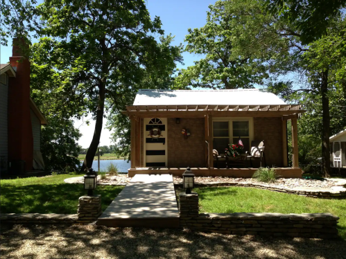 A charming cottage with a porch, surrounded by trees and a view of a lake, on a sunny day.