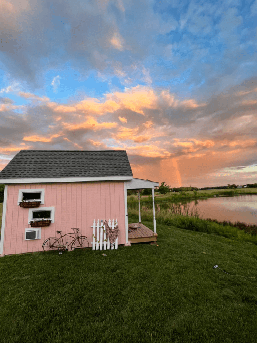 A pink shed by a pond, with a bicycle and flowers, under a colorful sunset sky with clouds.