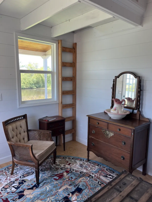 Cozy room featuring a vintage chair, dresser, mirror, and a decorative rug, with a window and wooden ladder.