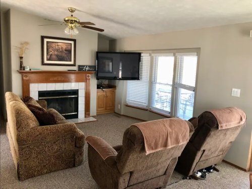 Cozy living room with a fireplace, two recliners, a sofa, and a large TV near windows.