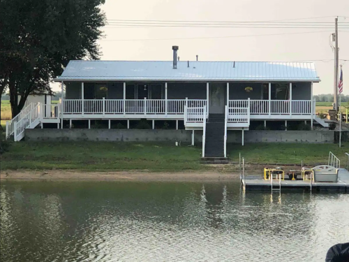 A gray house with a white porch and railing, situated by a calm body of water, surrounded by greenery.