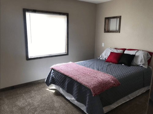 A cozy bedroom featuring a bed with gray and red bedding, a window with blinds, and a simple framed mirror.
