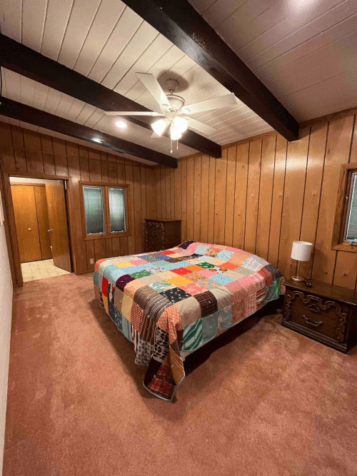 Cozy bedroom with a colorful quilt, wooden walls, ceiling beams, and a ceiling fan. Soft carpet and nightstands are visible.