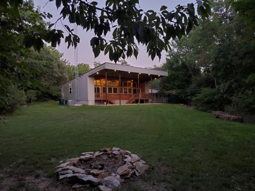 A modern house with a spacious deck, surrounded by trees and a grassy yard, with a stone fire pit in the foreground.