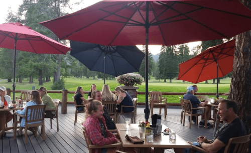 Outdoor dining area with wooden tables, umbrellas, and people enjoying meals in a scenic, tree-lined setting.