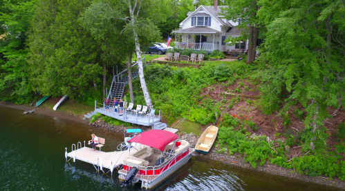 A lakeside house with a dock, boat, and seating area surrounded by trees and greenery.