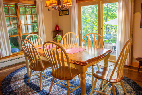A cozy dining room with a wooden table, six chairs, and large windows overlooking a green outdoor space.