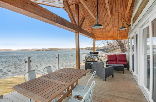 A spacious deck with a wooden table, chairs, and seating area overlooking a serene lake and distant hills.