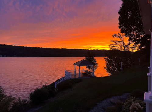 A vibrant sunset over a lake, with a gazebo silhouetted against the colorful sky and trees in the foreground.