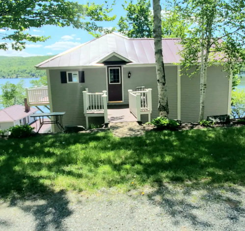 A charming cottage with a metal roof, surrounded by greenery and overlooking a lake on a sunny day.