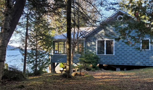 A cozy blue house nestled among trees, overlooking a serene lake on a sunny day.