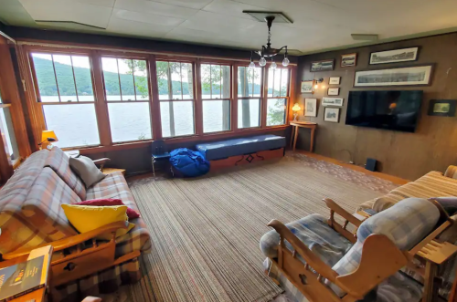 Cozy living room with large windows overlooking a lake, featuring comfortable seating and rustic decor.