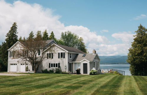 A serene lakeside house with a manicured lawn, surrounded by trees and mountains under a partly cloudy sky.