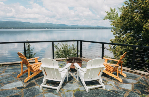 A serene lakeside view with four chairs around a fire pit on a stone patio, surrounded by trees and mountains.
