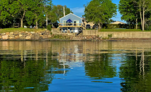 A blue house with a deck reflected in calm water, surrounded by trees and a clear blue sky.