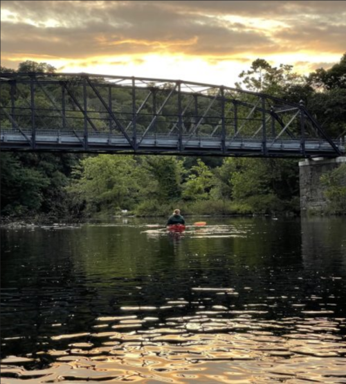 A person kayaking on a calm river under a bridge, surrounded by trees and a sunset sky.